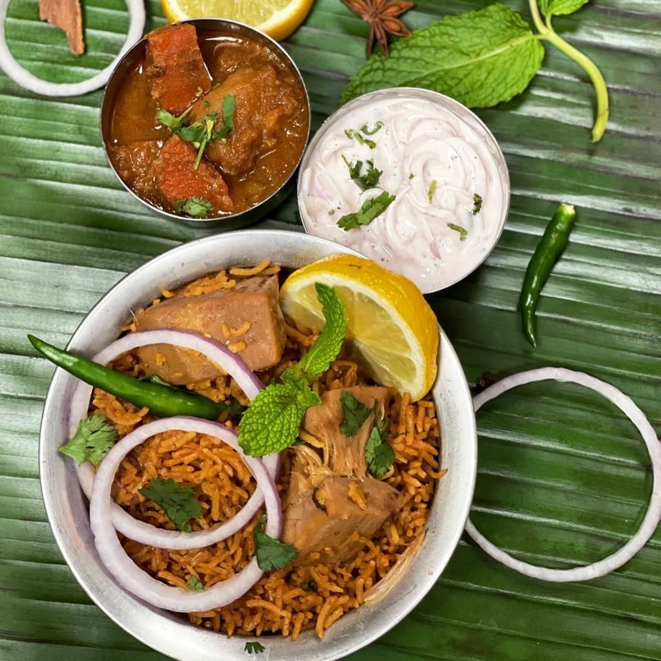 Indian meal featuring spiced rice, curry, raita, onion rings, lemon, green chilies, mint, and cilantro on banana leaves.