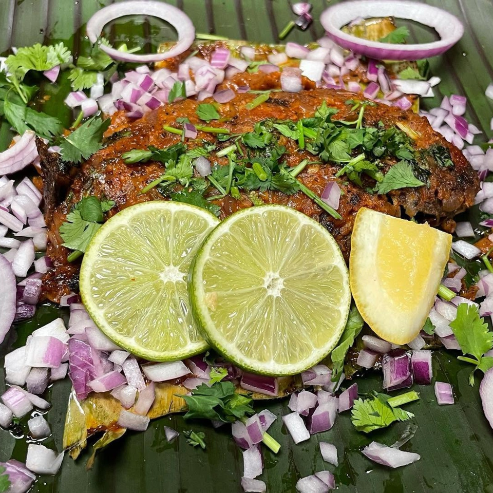 Grilled fish garnished with cilantro, chopped red onions, lime slices, and lemon wedges on a banana leaf.