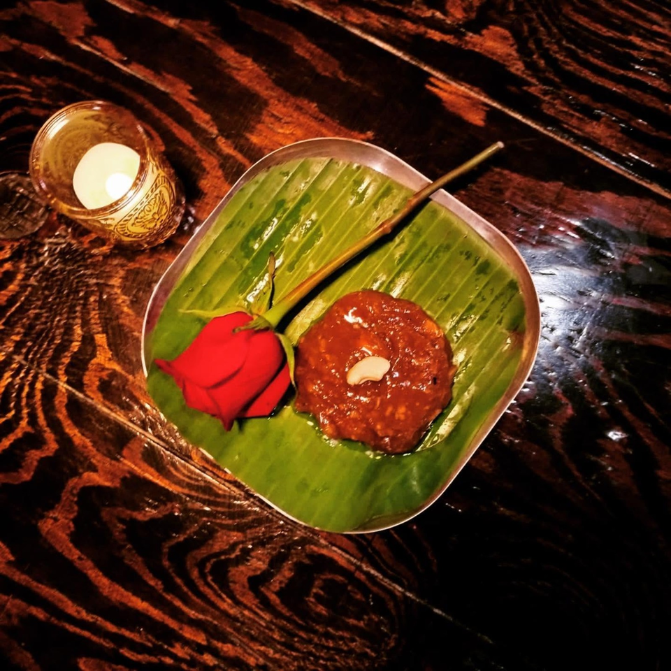 A banana-leaf-lined plate holds a red flower, saucy round dessert topped with a cashew, and a wooden skewer beside a candle.